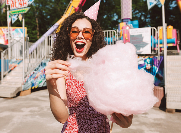 cheerful lady with dark curly hair sunglasses birthday cap standing with cotton candy hand happily looking camera while spending time amusement park